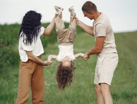 A joyful family enjoys outdoor play, lifting their child upside down in a green field.